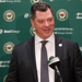 Bill Guerin stands in a black suit at a press conference. There is a green Minnesota Wild step-and-repeat behind him