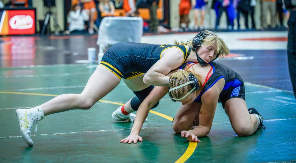 Image of the MImage of the Minneapolis Roosevelt High School Teddy wrestler on the mat during a meet. She’s got her opponent down on the ground using her weight as leverage as she wraps her up over the shoulder.