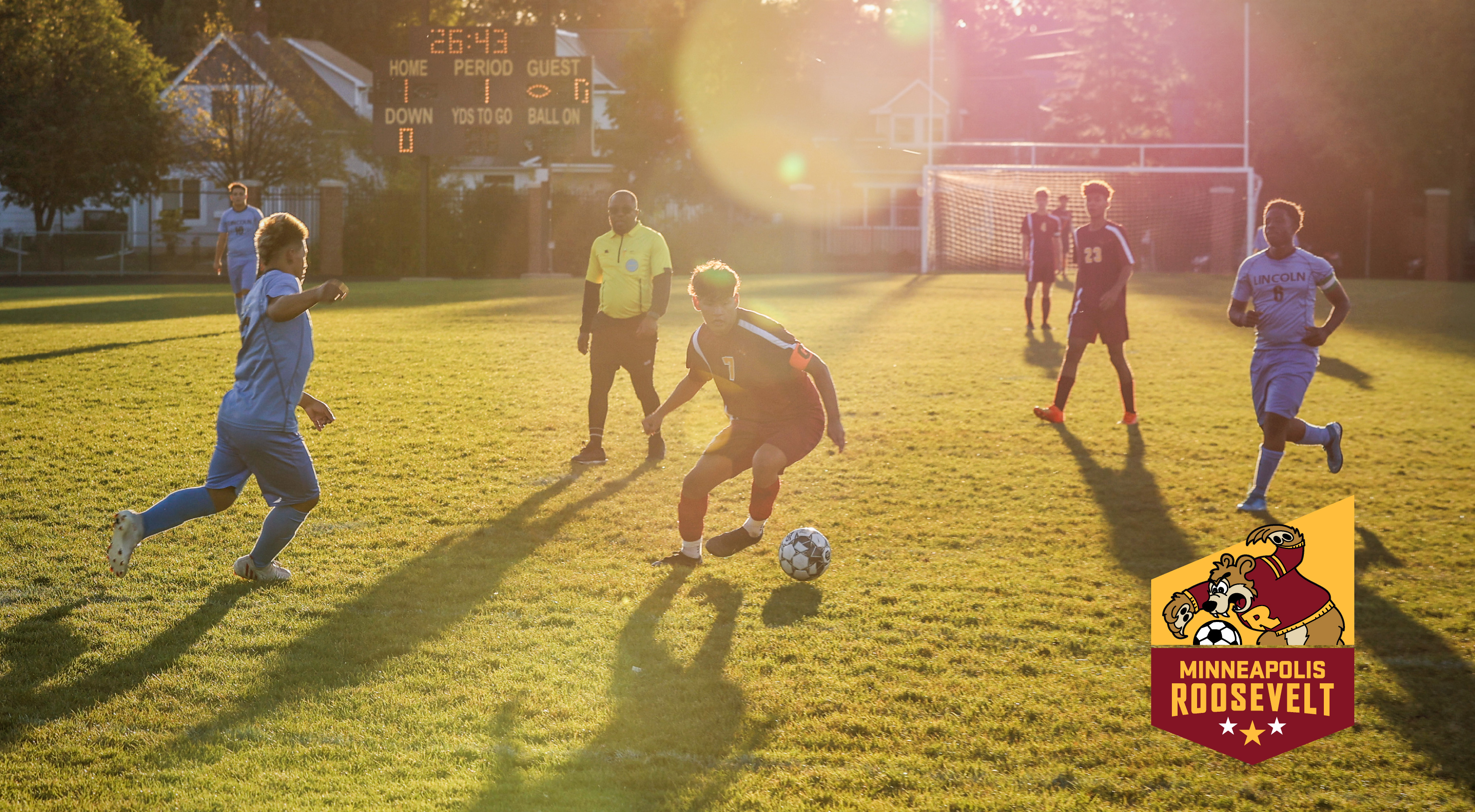 Image of Minneapolis Roosevelt High School Boys varsity soccer player advancing the ball upfield during an early evening game at Al Gowans Stadium in South Minneapolis, MN.. Superimposed over the image is a shield graphic with the illustrated “Teddy” logo