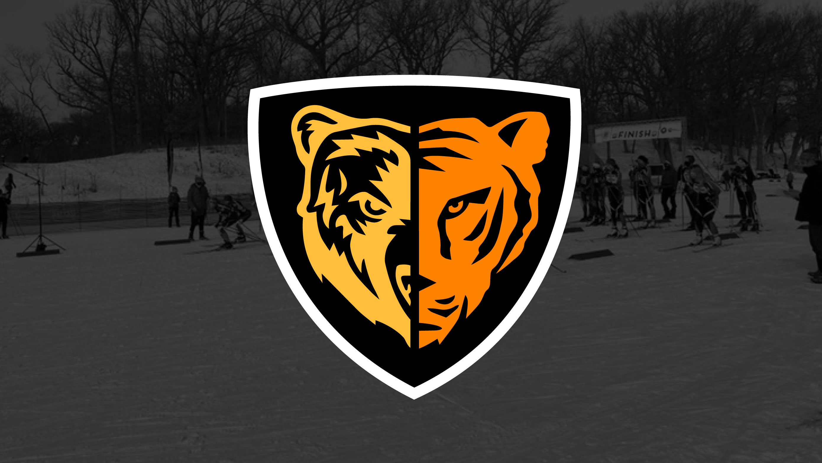 Image of a black-and-white photo of skiiers at the starting line at the begining of a meet. Superimposed on top is the TNT Logo made up of illustrations of half of a Bear’s Face on the left in Gold and half of a Tiger’s Face on the right. Both contained w