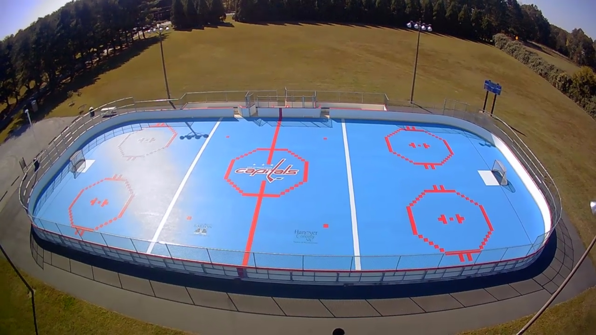 A roller hockey rink surrounded by white boards and chain link fencing.  The rink flooring is bright blue with red lines and a Washington Capitals logo in the center.  The sky is gray and cloudy.